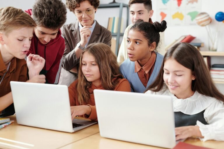 Students and a teacher gather around two laptops in a classroom, working together, with books and a map behind