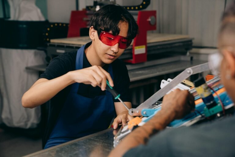 A young person wearing red safety glasses and a blue apron uses a screwdriver to work on a mechanical device at a workbench, while another person assists. Industrial equipment is visible in the background.