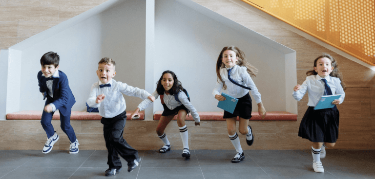 Five young children in school uniforms joyfully run indoors, some holding blue notebooks, with a wooden wall and yellow staircase in the background.