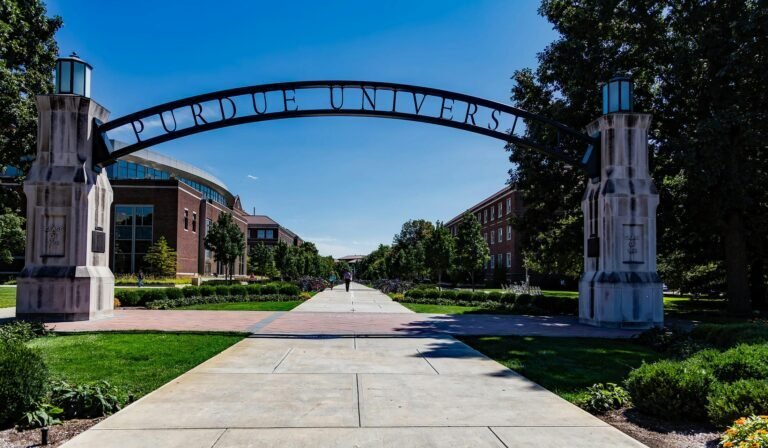 The Purdue University archway stands over a wide sidewalk lined with greenery and academic buildings on a sunny day with a clear blue sky.