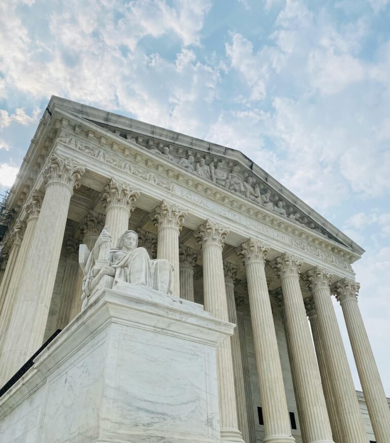 A low-angle view of the U.S. Supreme Court building, featuring tall columns and a marble statue in the foreground, with a partly cloudy sky overhead.