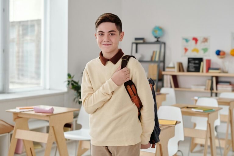 A smiling boy with short brown hair stands in a classroom, holding a backpack strap. The classroom has wooden desks, chairs, bookshelves, and educational posters on the walls. Daylight streams in through the window.
