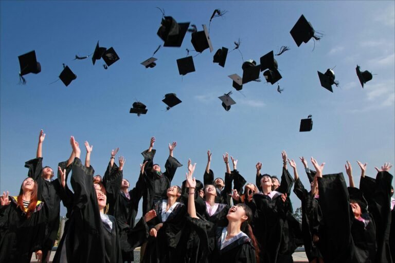 A group of graduates in black caps and gowns celebrate by throwing their caps into the air against a clear blue sky. They appear joyful and are looking upward with arms raised.