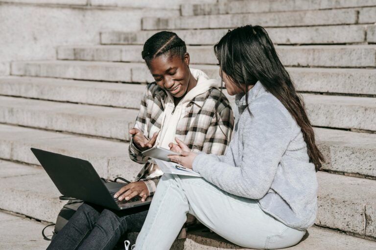 Two young people sit on outdoor steps, one with a laptop and the other holding a notebook. They are smiling and discussing something together, appearing engaged and collaborative.