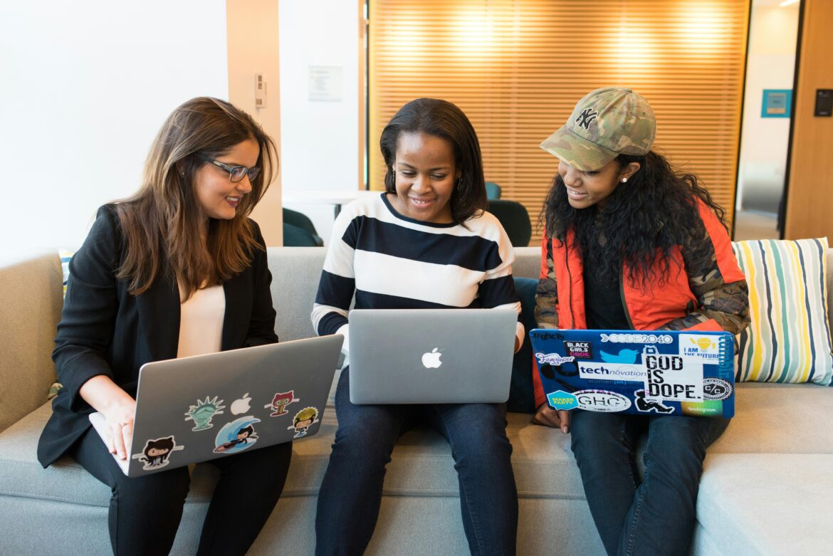 Three women sit on a couch using laptops, smiling and collaborating. Two laptops are decorated with stickers, and the background shows a modern office with wooden blinds and colorful cushions.
