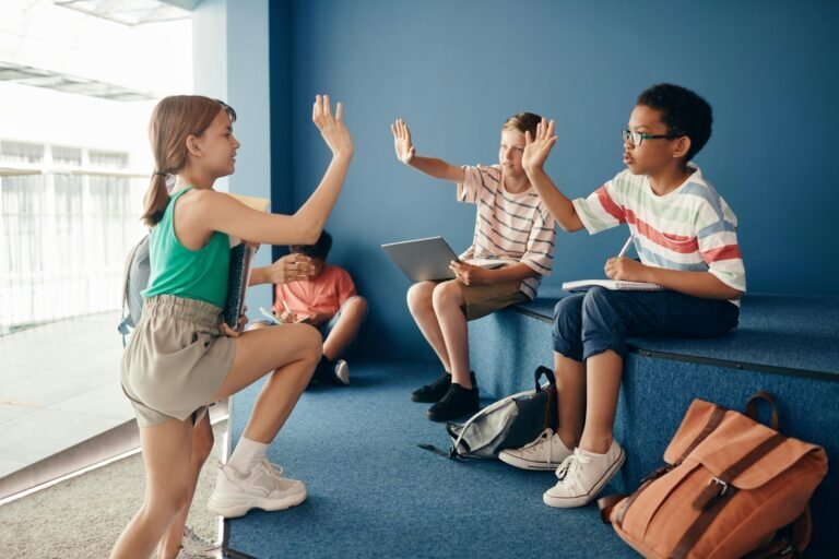 Four kids sit and talk in a modern indoor space. A girl and a boy give each other a high-five, while another boy uses a laptop and a fourth child sits in the background. Backpacks are on the floor nearby.