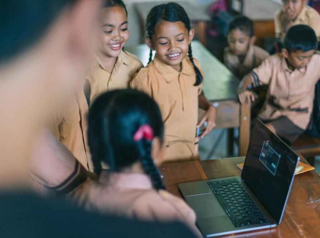 A group of young students in brown uniforms smile and gather around a laptop on a classroom desk while others interact in the background | edCircuit A group of young students in brown uniforms smile and gather around a laptop on a classroom desk while others interact in the background