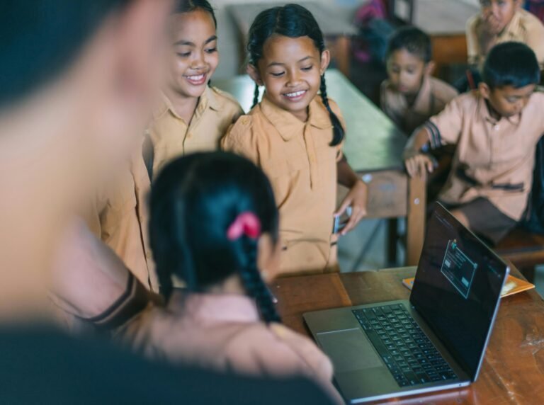 A group of young students in brown uniforms smile and gather around a laptop on a classroom desk, while others interact in the background.