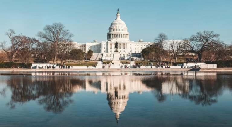 The United States Capitol building is seen in daylight, with clear blue skies and leafless trees. Its reflection appears in a calm pool of water in the foreground. People are walking and gathering near the steps.