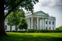 The White House in Washington DC is seen on a sunny day framed by large trees and surrounded by green grass and shrubs