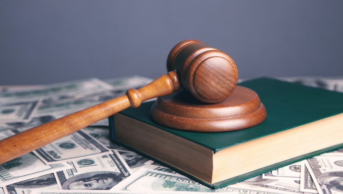 A wooden gavel resting on a green book surrounded by scattered dollar