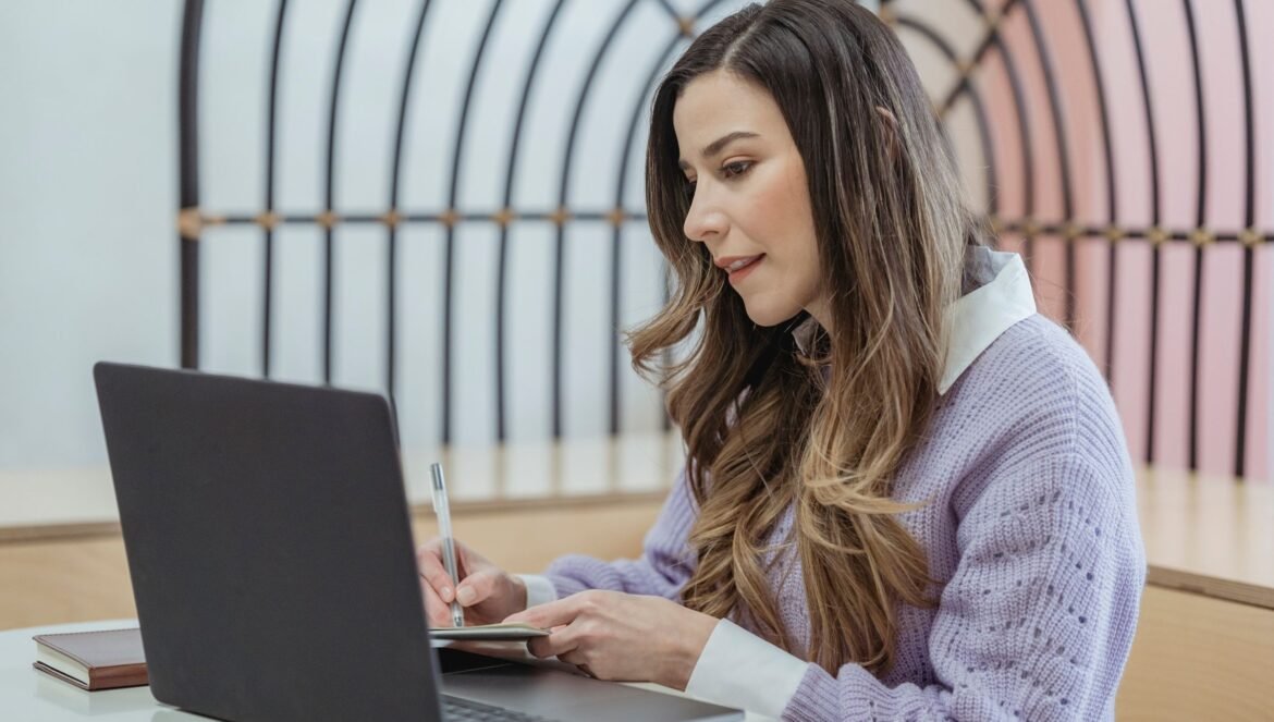 Focused female freelancer with wavy hair writing notes in planner whil
