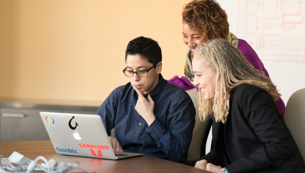 Three women beside table looking at MacBook