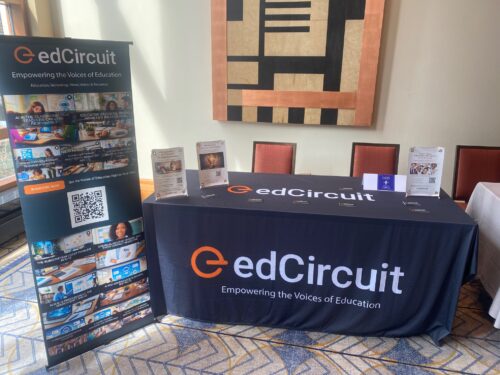 EdCircuit CoSN display table at an event, featuring a branded tablecloth, brochures, promotional materials, and a standing banner with images and the tagline Empowering the Voices of Education.