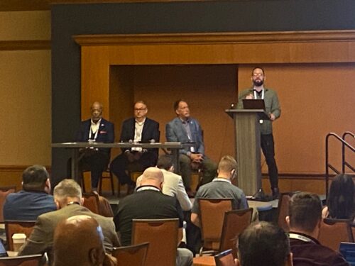 A speaker stands at a podium while three panelists, part of the CoSN conference, sit at a table onstage; audience members face the stage in a conference room.