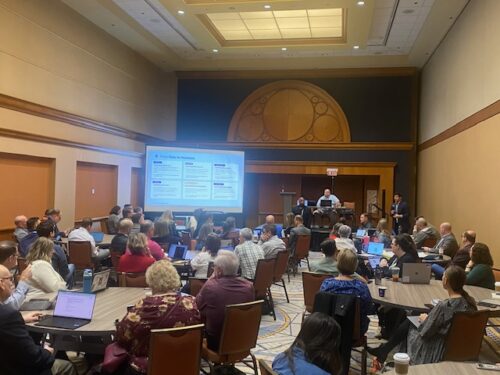 A conference room filled with people seated at tables, facing a presentation screen displaying a CoSN slide with text and graphics.