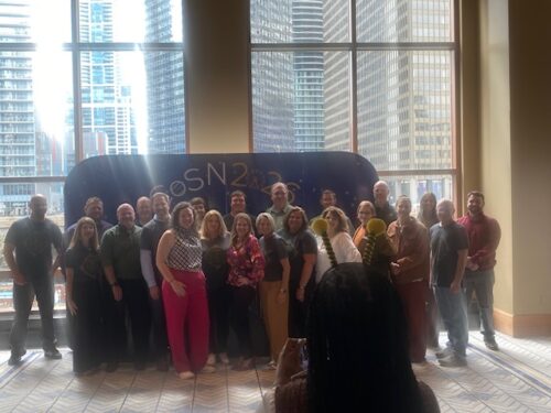 A group of people pose for a photo indoors in front of a sign reading “CoSN 2024,” with large windows and city buildings visible in the background.