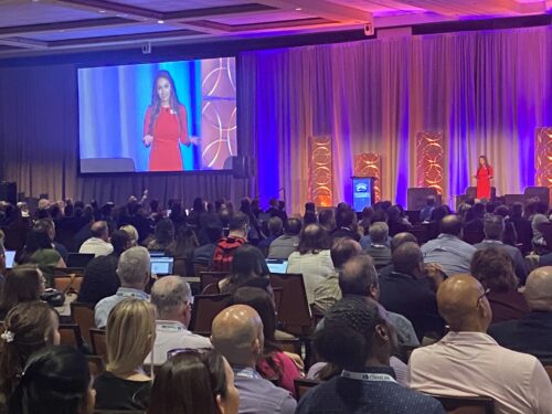 A woman in a red dress speaks on stage to a large seated audience at a CoSN conference, with her image projected on a screen beside her.