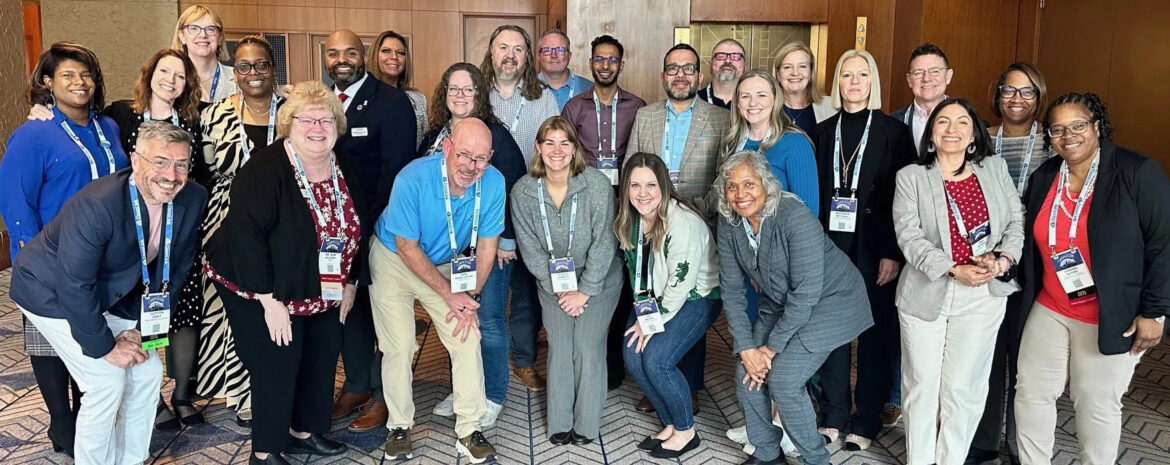 A group of 25 adults, both men and women, posing and smiling for a photo indoors at a conference or professional event.