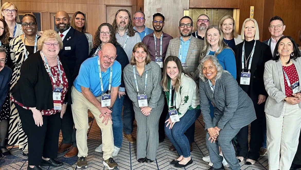 A group of 25 adults, both men and women, posing and smiling for a photo indoors at a conference or professional event.