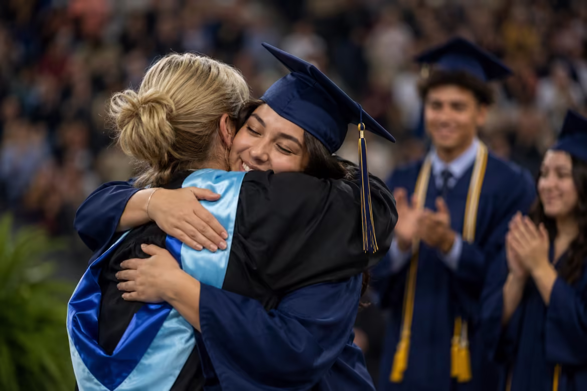 Graduate in a blue cap and gown hugs a smiling person at a graduation ceremony as classmates applaud in the background of a crowded arena.