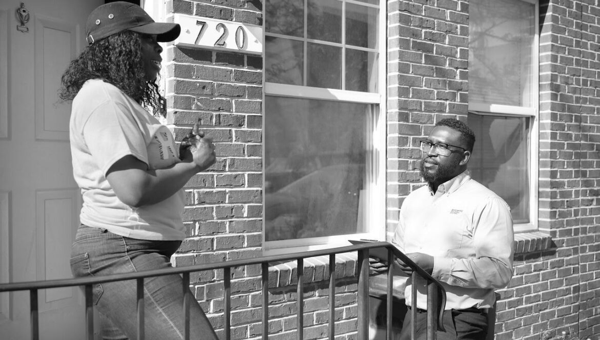 Two people stand outside a brick townhouse a woman wearing a cap talks to a man holding a clipboard