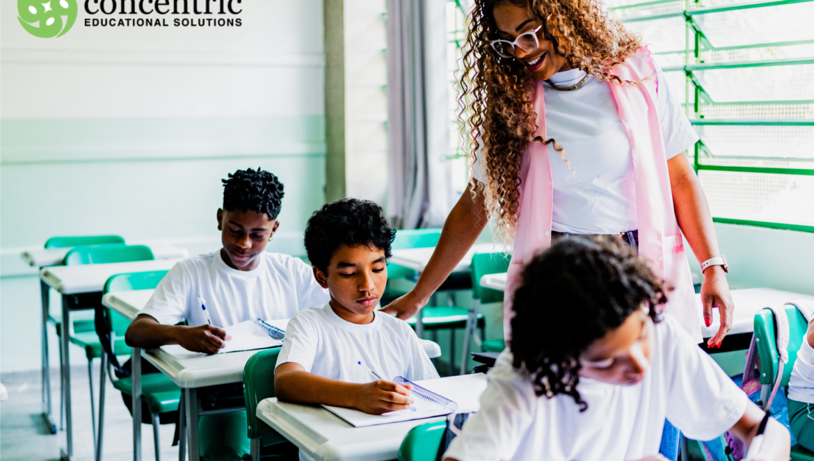 Teacher in a pink vest helps students at desks in a bright classroom with concentric educational solutions logo on the left