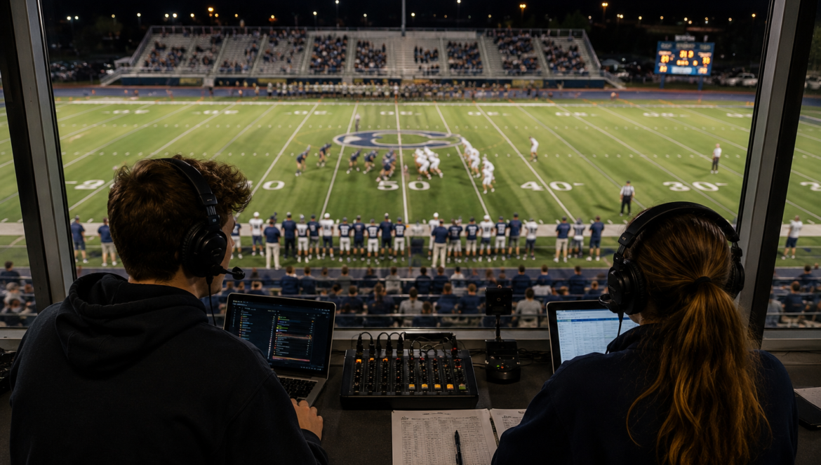 Two sports commentators wearing headsets in a broadcast booth | edCircuit Two sports commentators wearing headsets in a broadcast booth overlook a nighttime football game on a lit field