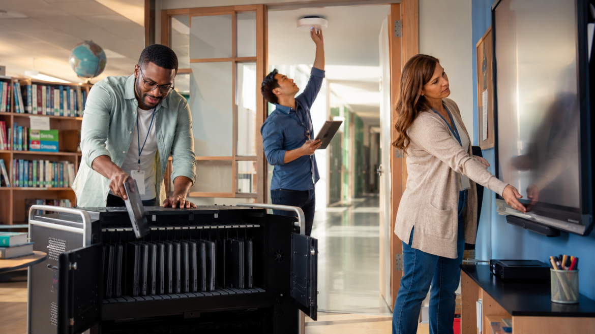 Three adults in a library: man loading a large printer, another man holding a tablet and reaching the ceiling sensor, woman using a touchscreen board.