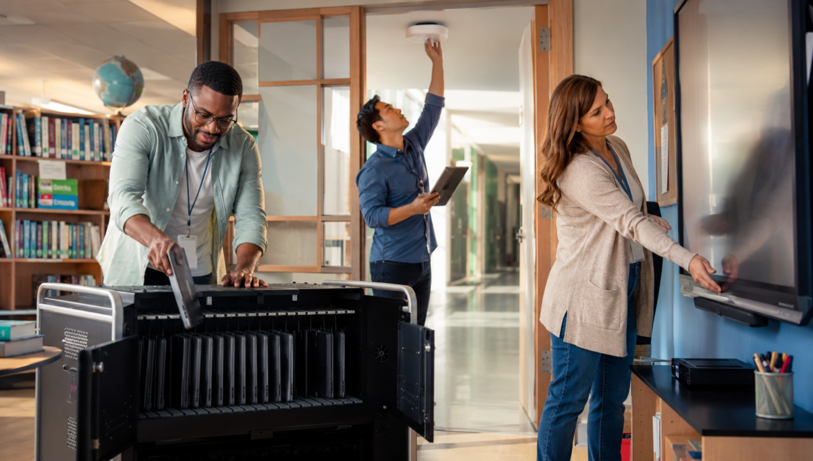 Three adults in a library man loading a large printer another man holding a tablet and reaching the ceiling sensor woman using a touchscreen board