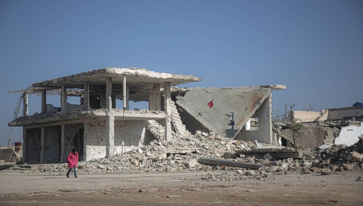 A child in a pink coat walks near a war torn building with an artwork on the wall