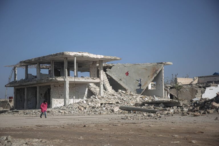 A child in a pink coat walks near a war-torn building with an artwork on the wall