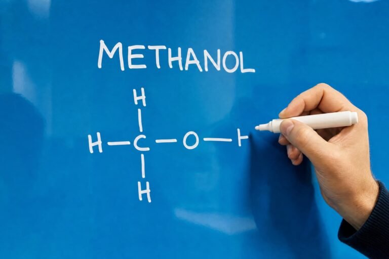 A hand drawing the chemical structure of methanol with the word METHANOL above it in white marker on a bright blue glass board