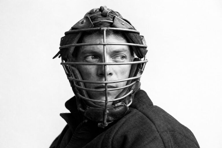 A man in a dark jacket wearing an old metal baseball catcher's mask, looking to the side, plain light background, dramatic studio light
