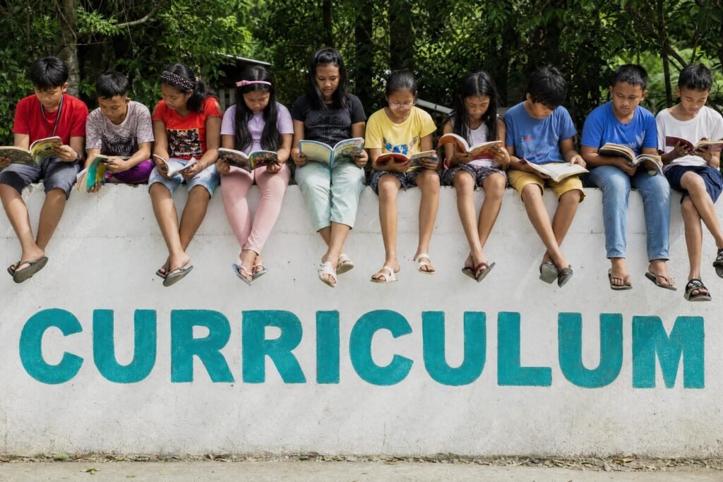 A row of children sitting on top of a white wall reading books with the word CURRICULUM painted on the wall below them in teal letters | edCircuit A row of children sitting on top of a white wall reading books with the word CURRICULUM painted on the wall below them in teal letters