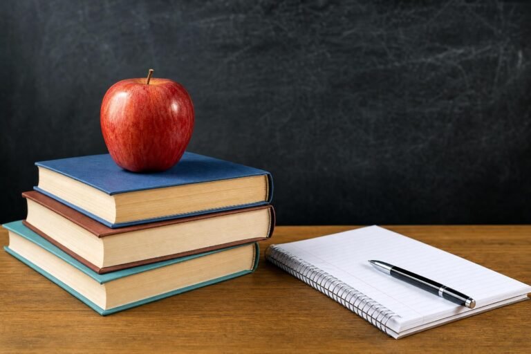 A stack of books with a red apple on top beside an open notebook and pen, dark chalkboard behind