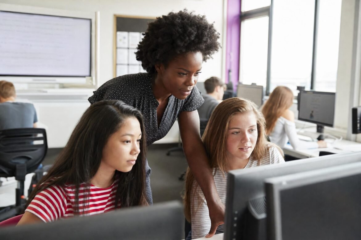 A teacher leaning over to help two teenage girls working at desktop computers in a bright computer classroom