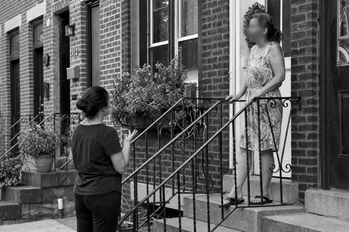 A woman on the front steps of a brick row house talking with a neighbor standing on the stoop next door, documentary style, faces not identifiable