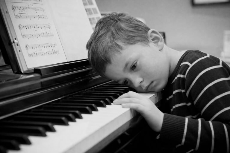 A young boy in a striped sweater resting his head on the keys of a piano, looking sideways, sheet music on the stand