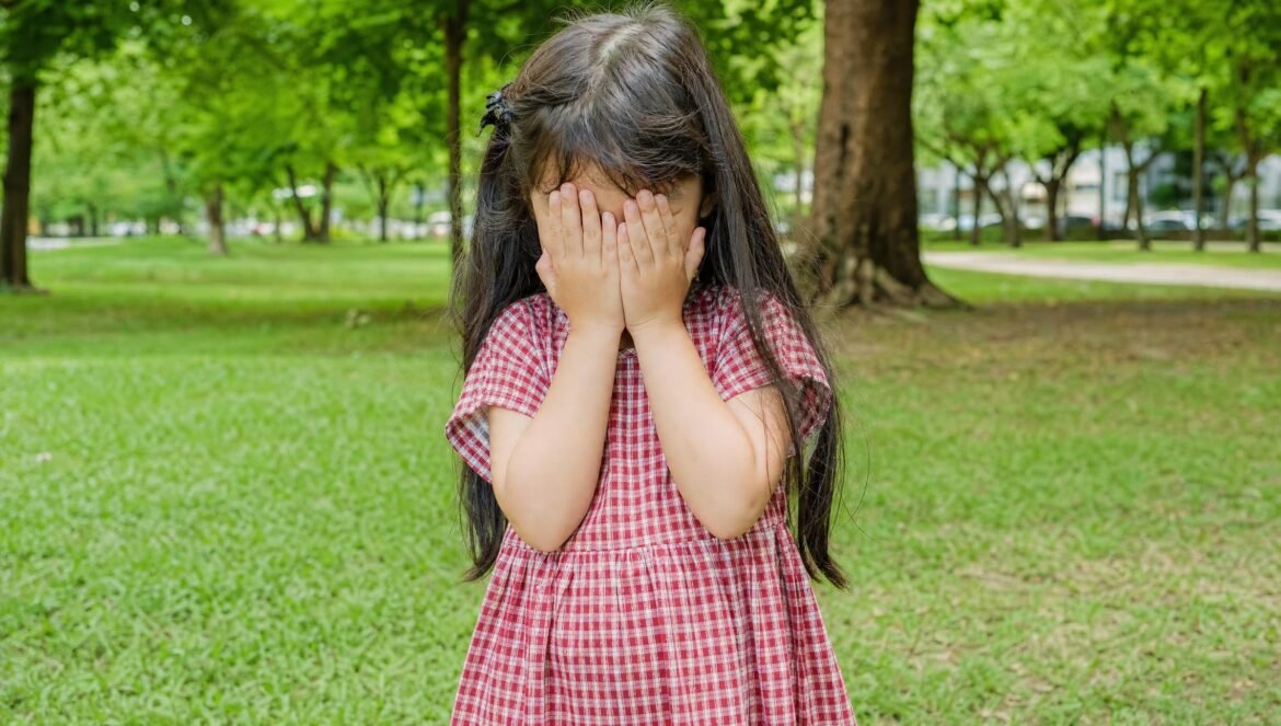 A young girl with long dark hair in a red checked dress standing in a green park covering her face with both hands shy daylight | edCircuit A young girl with long dark hair in a red checked dress standing in a green park covering her face with both hands shy daylight