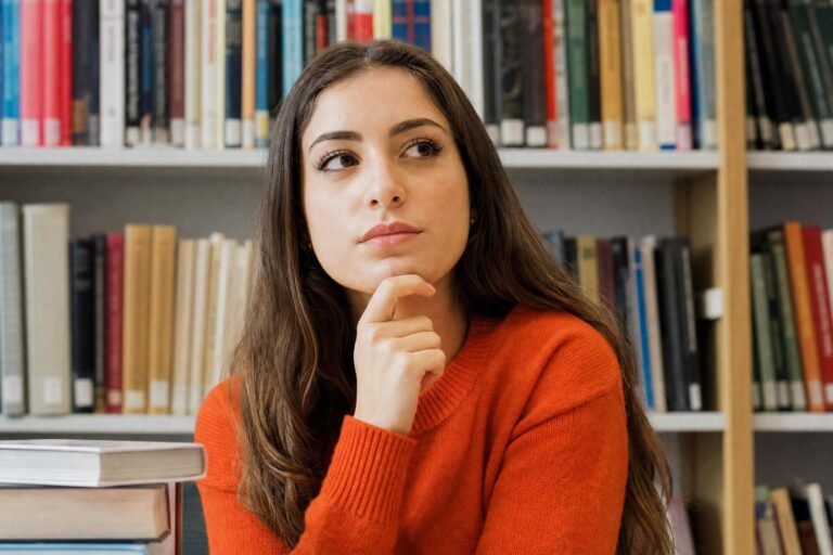 A young woman in an orange sweater with her hand on her chin, thinking, in front of library bookshelves