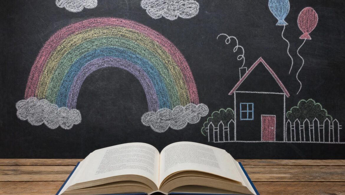 An open book on a wooden table in front of a dark chalkboard covered with chalk drawings of a rainbow a house clouds and balloons