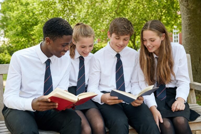 Four teenage students in school uniforms sitting on a wooden bench outdoors reading books together, summer trees behind