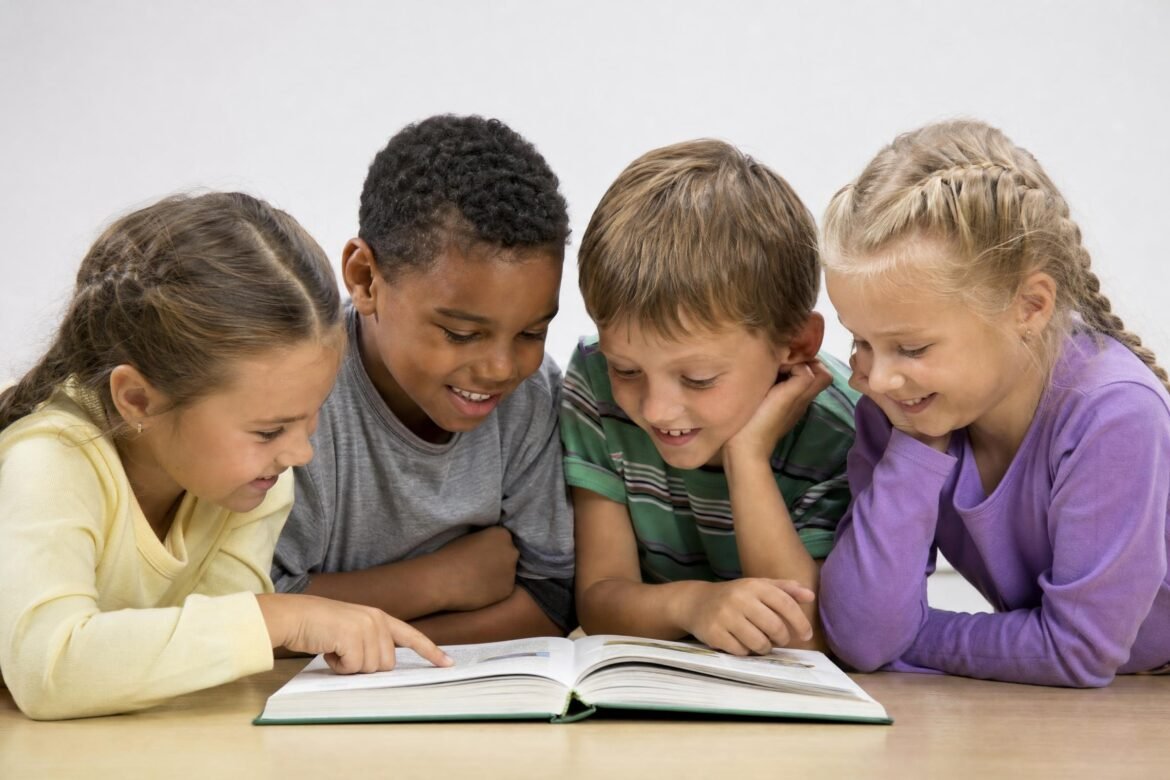 Four young children leaning over a table reading a book together, smiling, plain light background