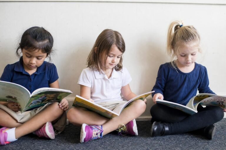 Three young girls in simple school clothes sitting side by side on a classroom floor against a white wall, each reading an open picture book, soft natural light