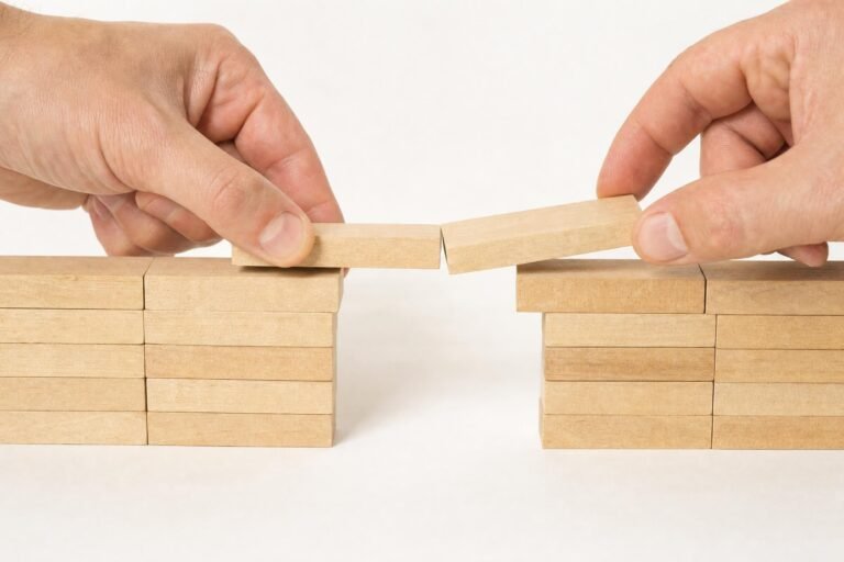 Two hands each placing the last wooden block to complete a small bridge between two stacks of blocks, white background