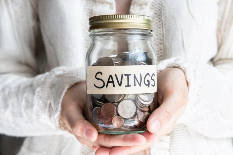 Two hands holding a glass jar full of coins with a handwritten paper label reading SAVINGS, soft light