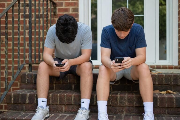 Two teenage boys sitting on outdoor brick steps looking down at their phones, one with his head bowed, daylight