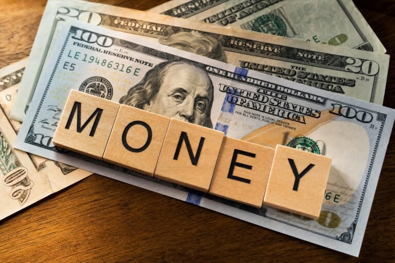 Wooden letter tiles spelling MONEY lying across a spread of US dollar bills on a table, warm side light, sharp detail