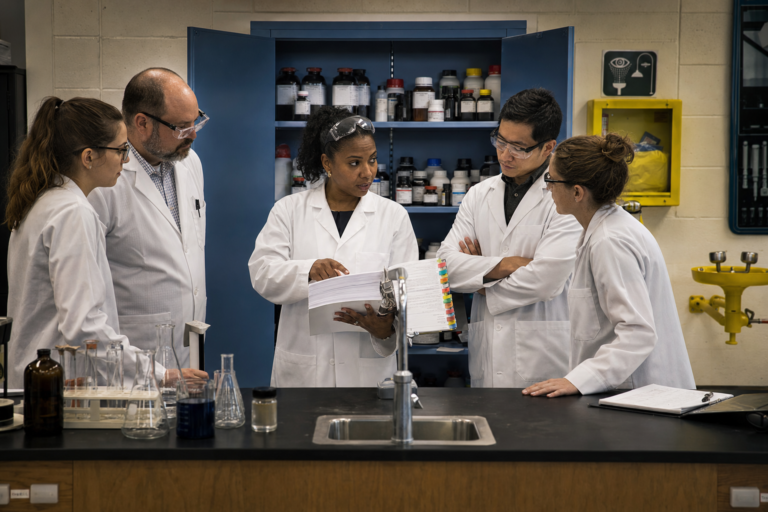 Chemical hygiene plan in science education: a chemical hygiene officer walks science teachers in lab coats through the tabbed plan binder beside an organized chemical storage cabinet and eyewash station